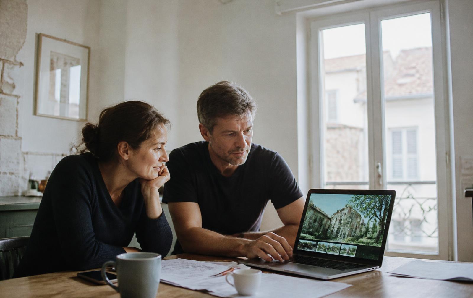 A couple reviewing a French property listing on a laptop at their kitchen table