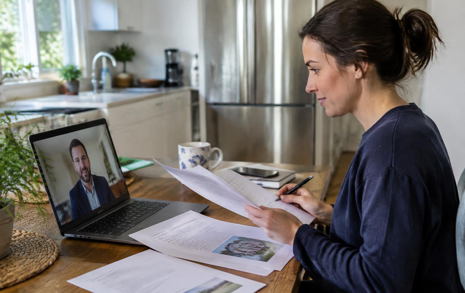 Young woman on a video call reviewing French property documents at her kitchen table